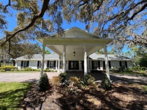 A white single-story house with a large covered entrance supported by tall columns, under a blue sky. The house is surrounded by lush greenery, trees with Spanish moss, and a brick driveway with colorful shrubs in the foreground.